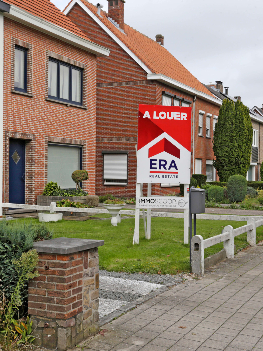 Il y a un grand panneau en V rouge dans le jardin avant d'une maison qui est mise en vente par ERA.